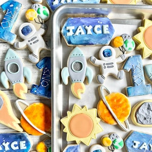 May include: A collection of space-themed sugar cookies featuring astronauts, rockets, planets, suns, and the number one, with some cookies personalized with the name JAYCE in white lettering against a blue and purple galaxy-inspired background.