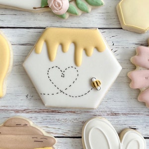 May include: A hexagonal sugar biscuit decorated with white icing, featuring a yellow honey drip pattern at the top, a black dashed line forming a heart shape in the centre, and a small three-dimensional bee figure resting on the right side.