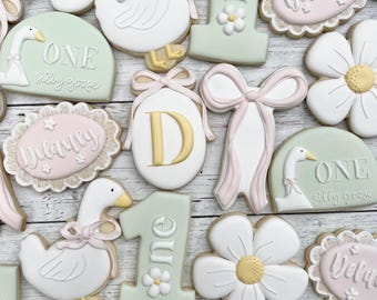 Può includere: Una collezione di biscotti decorati con margherite bianche dal centro giallo, oche bianche con fiocchi rosa, numeri uno verde chiaro con piccoli fiori, targhe ovali con il nome Delaney e fiocchi rosa pallido su una superficie bianca.
