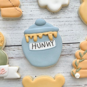 May include: A sugar cookie shaped like a blue honey pot with a lid, featuring yellow icing dripping from the top rim and a white rectangular label in the center with the word HUNNY written in black icing, surrounded by other decorative cookies.