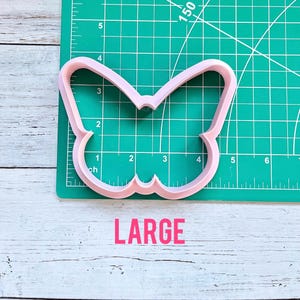 May include: A light pink butterfly-shaped cookie cutter positioned on a green cutting mat with a grid. The cookie cutter spans approximately 4.5 inches in width and 3 inches in height. The word LARGE is printed in pink below the cutter.