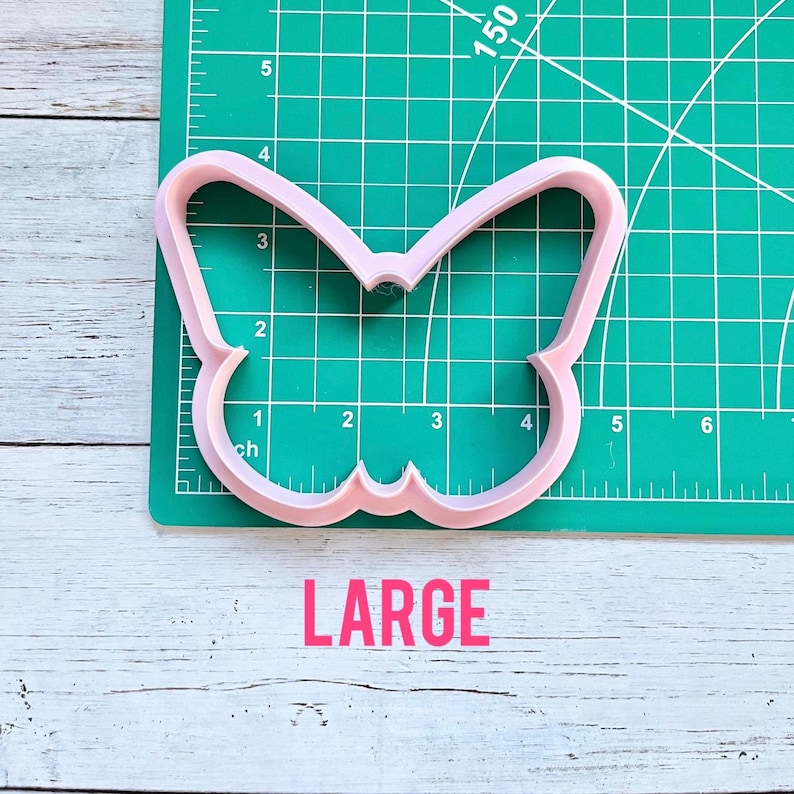 May include: A light pink butterfly-shaped cookie cutter positioned on a green cutting mat with a grid. The cookie cutter spans approximately 4.5 inches in width and 3 inches in height. The word LARGE is printed in pink below the cutter.