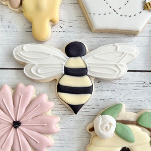 May include: A sugar cookie shaped like a bee with black and pale yellow stripes on the body and white icing wings featuring piped details. The bee cookie is surrounded by other cookies, including a pink flower and a white honeycomb shape.