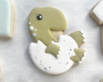 Puede incluir: Una galleta con forma de bebé dinosaurio saliendo de un huevo blanco. El dinosaurio es de color verde salvia con un ojo negro y manchas azul claro. La cáscara de huevo blanca presenta pequeñas motas negras y bordes festoneados blancos.