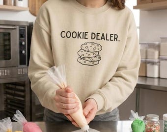 Può includere: Una felpa girocollo beige con la scritta nera "COOKIE DEALER." sopra un'illustrazione a linee di una pila di tre biscotti con gocce di cioccolato. Il capo presenta maniche lunghe e bordi a coste su scollo, polsini e orlo.