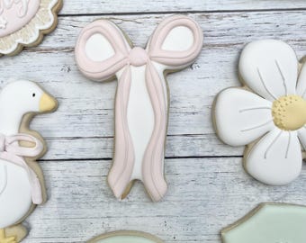 Può includere: Una collezione di biscotti decorati con un fiocco rosa pallido, un'oca bianca con un nastro rosa, una margherita bianca con il centro giallo, un numero uno verde pallido con un fiore bianco e un biscotto rotondo verde pallido con il testo ONE silly goose.