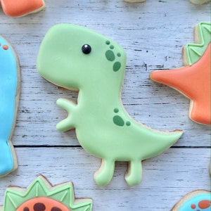 May include: A light green dinosaur-shaped sugar cookie decorated with smooth royal icing. The cookie features a small black dot for an eye and darker green spots along the neck and back, with a simple outline of a T-Rex silhouette.
