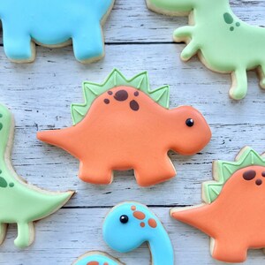 May include: A collection of dinosaur-shaped sugar cookies decorated with smooth royal icing. The cookies feature various species in shades of orange, light blue, and pale green, with detailed icing accents including spots and spikes.