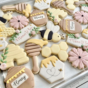 May include: A collection of decorated sugar cookies featuring bee-themed shapes including honey pots labeled Honey, honey dippers, honeycomb patterns, bees, pink flowers, and rectangular plaques with the script text Mama to bee.