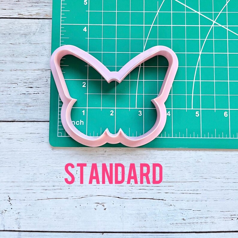 May include: A light pink butterfly-shaped cookie cutter positioned on a green cutting mat with a white grid. The cutter measures approximately 3.5 inches in width and 2.5 inches in height. The word STANDARD is printed in pink below the mat.