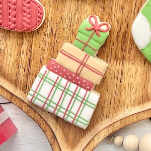 May include: A stack of four decorated sugar cookies shaped like gift boxes. The top box is green with a red bow, the second is tan with red lines, the third is red with white dots, and the bottom box is white with a green and red plaid pattern.