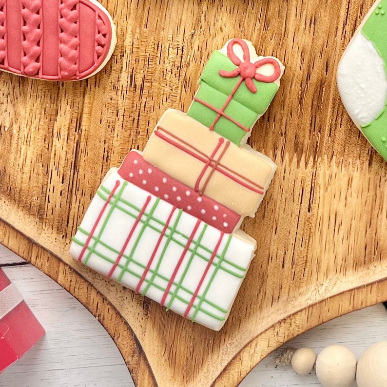 May include: A stack of four decorated sugar cookies shaped like gift boxes. The top box is green with a red bow, the second is tan with red lines, the third is red with white dots, and the bottom box is white with a green and red plaid pattern.