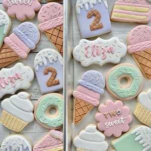 May include: A collection of decorated sugar cookies shaped like ice cream cones, cupcakes, donuts, cake slices, and popsicles. Some cookies feature the text "TWO sweet" or "Eliza" and the number "2" in pastel icing with colorful sprinkles.