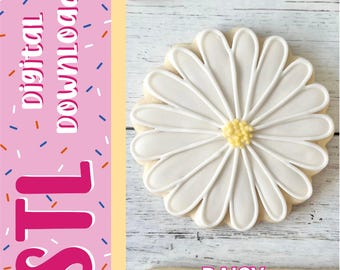 Può includere: Un biscotto a forma di margherita con petali di glassa bianca e centro giallo, accanto a una barra laterale rosa con il testo digital download, STL e DAISY. Il biscotto è posizionato su una superficie in legno chiaro con venature.