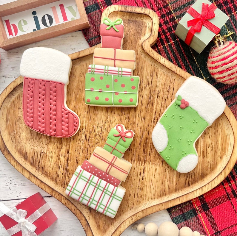 May include: Four decorated sugar cookies shaped as two Christmas stockings and two stacks of gift boxes. The stockings are red with a white cuff and green with a white cuff. The gift stacks feature various colors including red, green, and tan.