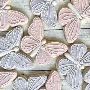 May include: A collection of butterfly-shaped sugar cookies decorated with smooth royal icing in soft pastel shades of lavender and dusty pink. Each cookie features intricate white piped line details and small dots on the wings.