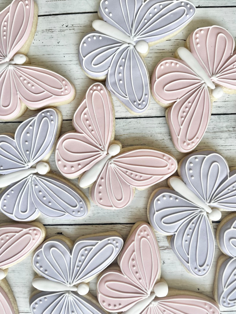 May include: A collection of butterfly-shaped sugar cookies decorated with smooth royal icing in soft pastel shades of lavender and dusty pink. Each cookie features intricate white piped line details and small dots on the wings.