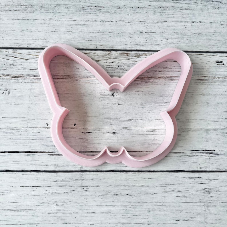 May include: A light pink butterfly-shaped cookie cutter with a hollow center and defined wing outlines. The tool features a smooth, rigid plastic construction with a consistent depth, designed for cutting shapes out of dough or other materials.