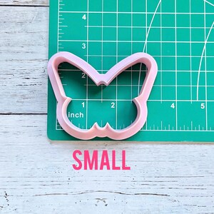 May include: A light pink butterfly-shaped cookie cutter positioned on a green cutting mat with white grid lines. The cutter measures approximately 2.5 inches in width and 2 inches in height. The word SMALL is printed in pink below the cutter.