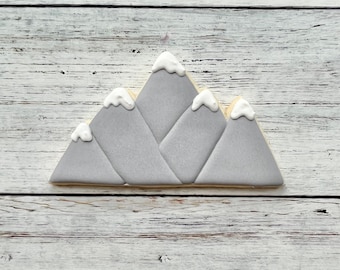 Puede incluir: Una galleta con forma de montaña decorada con glaseado gris liso y detalles de glaseado blanco en los picos para representar la nieve. La galleta presenta cuatro secciones triangulares distintas superpuestas para crear una cordillera con una base plana.