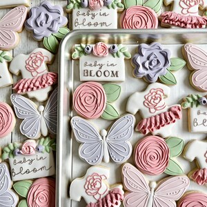 May include: A collection of decorated sugar cookies featuring light pink and lavender butterflies, pink roses with green leaves, and white onesies with pink ruffles. Several rectangular cookies display the text "baby in BLOOM" in dark lettering.
