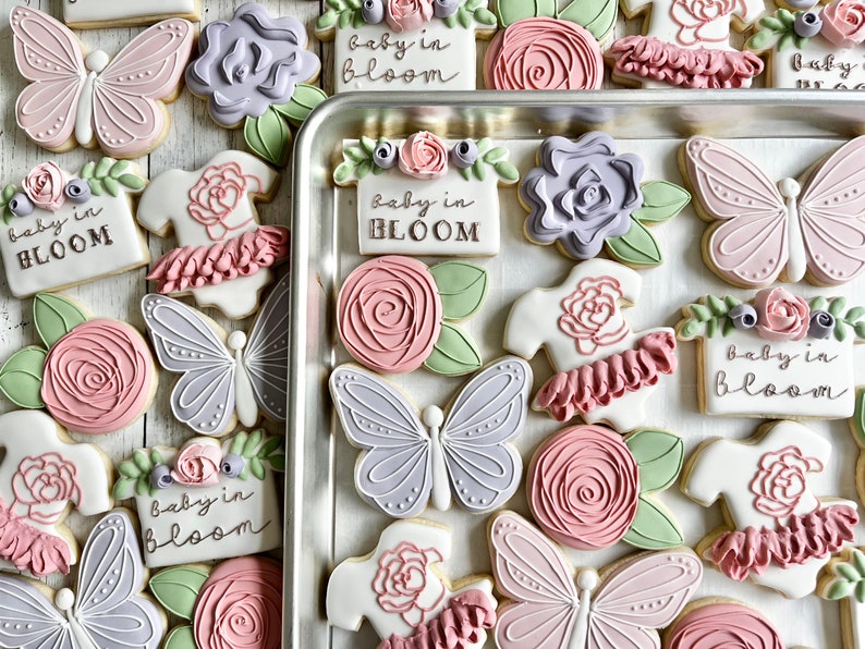 May include: A collection of decorated sugar cookies featuring light pink and lavender butterflies, pink roses with green leaves, and white onesies with pink ruffles. Several rectangular cookies display the text "baby in BLOOM" in dark lettering.