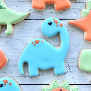 May include: A light blue dinosaur-shaped sugar cookie with smooth icing, featuring orange spots on its back and neck, and a single small black dot for an eye. The cookie is surrounded by other dinosaur-shaped cookies in shades of green and orange.