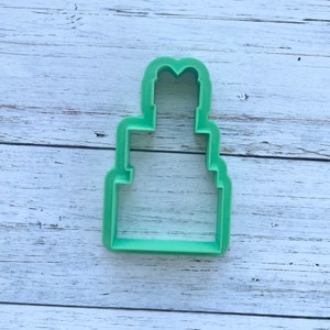 May include: A light green plastic cookie cutter shaped like a three-tiered cake. The design features a wide rectangular base, a narrower middle section, and a smaller top tier with a rounded, heart-like peak at the very top of the structure.