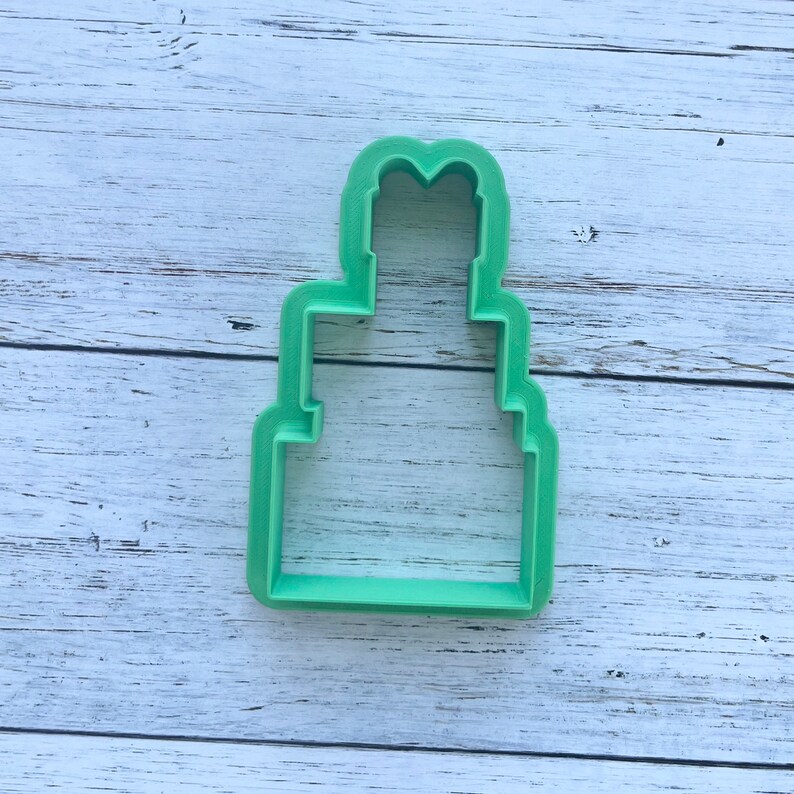 May include: A light green plastic cookie cutter shaped like a three-tiered cake. The design features a wide rectangular base, a narrower middle section, and a smaller top tier with a rounded, heart-like peak at the very top of the structure.