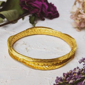May include: A gold-toned bangle bracelet with a textured, hammered finish. The bracelet is thick and has a slightly curved shape.
