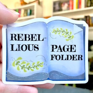 Puede incluir: Una pegatina azul y blanca con un diseño de libro. La pegatina dice "Rebellious Page Folder" con enredaderas verdes en la portada.