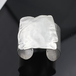 May include: A silver cuff bracelet with a textured, square-shaped design. The bracelet is open on one side and has a smooth, polished finish.