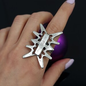 May include: A silver ring with a geometric design resembling a starburst or flower. The ring is worn on a finger with a purple background.