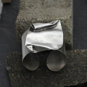 Puede incluir: Un brazalete de plata con un diseño texturizado y doblado. El brazalete está abierto y listo para ser usado.