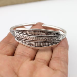 Op de afbeelding: Een zilveren armband met een geometrisch patroon. De armband is open en heeft een gebogen vorm.