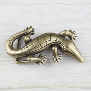 May include: A gold-toned metal belt buckle shaped like an alligator. The buckle has a detailed design with scales and claws.