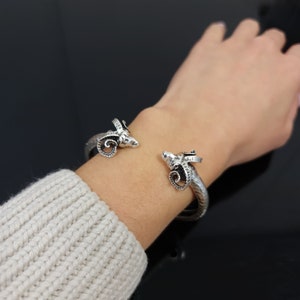 May include: A silver bracelet with ram's head accents. The bracelet is open and has a hammered texture.