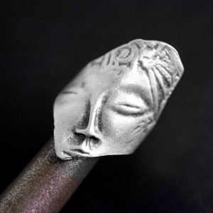 May include: A silver ring with a detailed face design. The face has closed eyes and a small mouth. The ring is on a brown surface.