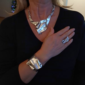 Könnte beinhalten: Ein silbernes Halsketten- und Armbandset mit geometrischem Design. Die Halskette ist ein Choker-Stil mit einem großen, gefalteten Stück in der Mitte. Das Armband ist ein breiter Manschettenarmreif mit einem ähnlichen gefalteten Design.