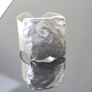 Op de afbeelding: Een zilveren armband met een getextureerd, gehamerd, vierkant ontwerp. De armband is aan &eacute;&eacute;n kant open en heeft een gladde, gepolijste afwerking.
