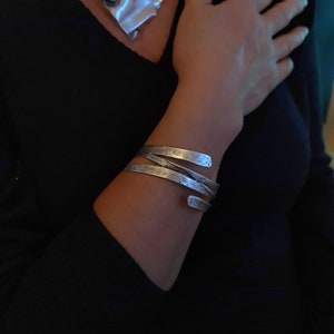 Könnte beinhalten: Ein silbernes Armband mit gehämmerter Oberfläche. Das Armband besteht aus drei Metallbändern, die miteinander verdreht sind.