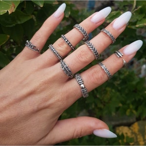 Può includere: Un set di nove anelli d'argento con vari design, tra cui una foglia, una corona e motivi geometrici. Gli anelli sono indossati su una mano con unghie lunghe e bianche.