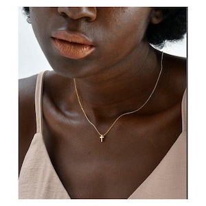 Pode incluir: Um delicado colar dourado com um pequeno pingente em forma de cruz. O colar é usado no pescoço de uma pessoa, contra um fundo de um top de tom neutro. O pingente de cruz é centralizado e a corrente é fina.