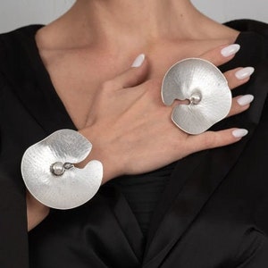 Könnte beinhalten: Silberfarbenes Schmuckset mit einem Armreif und einem Ring. Das Armband umschließt das Handgelenk, und der Ring sitzt am Finger. Beide Teile haben ein strukturiertes, organisches Design mit einem zentralen, abgerundeten Akzent.