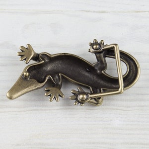 May include: A bronze-colored belt buckle shaped like a gecko lizard. The buckle is detailed with textured scales and has a hinged design.