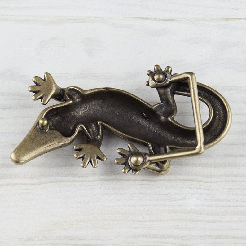 May include: A bronze-colored belt buckle shaped like a gecko lizard. The buckle is detailed with textured scales and has a hinged design.