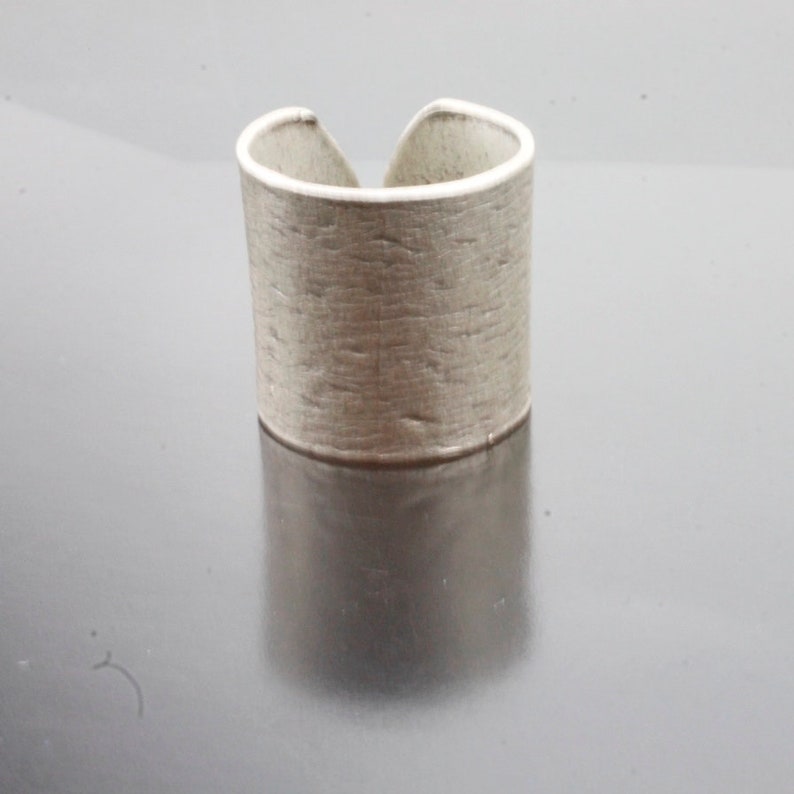 Puede incluir: Un anillo de plata con un acabado texturizado y martillado. El anillo es ancho y tiene una forma ligeramente redondeada.