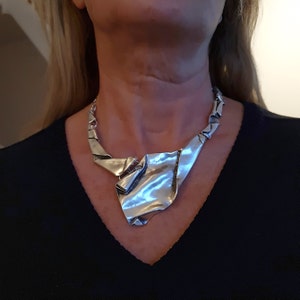 Puede incluir: Un collar de declaración plateado con un diseño grande, abstracto y doblado. El collar lo lleva una persona con una camisa negra.