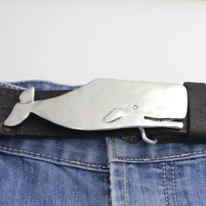 Może przedstawiać: Srebrna klamra do paska w kształcie wieloryba przymocowana do czarnego skórzanego paska. Klamra ma szczegółowe płetwy i ogon, na tle niebieskich dżinsów.