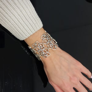 Könnte beinhalten: Ein silbernes Armband mit einem filigranen, komplizierten Design aus sich drehenden, miteinander verbundenen Schleifen. Das Armband ist breit und hat eine leicht gebogene Form.